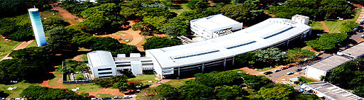 Imagem aérea de um edifício alongado e curvo, de cor clara, cercado por áreas verdes e árvores.Ao redor, há caminhos, ruas e outras construções, compondo um ambiente que lembra um campus ou complexo institucional.